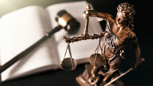 A statute of a blindfolded woman holding up the scales of justice sitting on a table next to an open law book with a judge's gavel sitting on top of it.